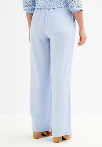 Pantalones de pierna ancha de color azul claro, confeccionados en un tejido suave, con una cinturilla elástica y un bolsillo trasero, combinados con zapatos de tacón de madera.