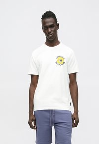 Valkoinen puuvillainen t-paita, jossa keltamusta auringonkukkakuvio ja teksti "SUNNIES POOL BAR". Päällä violetit shortsit, lyhyet hihat.