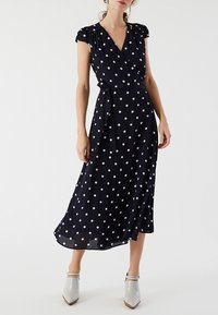 Femme portant une robe midi bleu marine à pois blancs, manches courtes, col en V, ceinture à la taille, et chaussures blanches à bout pointu.