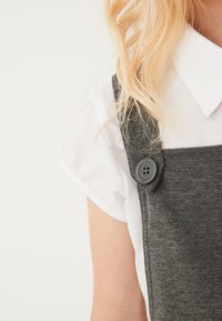 Szare bawełniane ogrodniczki o teksturowanym wykończeniu, z czarnym guzikiem, zestawione z białą bluzką z kołnierzykiem. Zbliżenie na odzież.