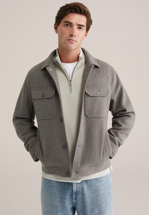 Grijs wollen mix buitenjas met twee borstzakken en knoopsluiting, gedragen over een lichtgroene top met rits, gecombineerd met lichtblauwe jeans.