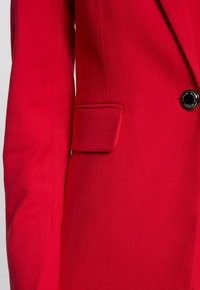 Blazer rosso con vestibilità strutturata, chiusura a un bottone e dettaglio delle tasche a filetto. Il tessuto ha una finitura liscia e leggermente texturizzata.