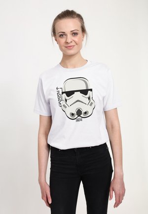 Jeune femme aux cheveux clairs portant un t-shirt blanc avec un graphique de casque de Stormtrooper noir et gris, et un pantalon noir, sur un fond uni.