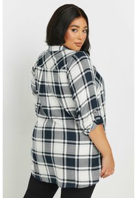 Camisa de cuadros con un patrón en tonos azul marino, blanco y gris, mangas largas remangadas hasta los codos y detalle de yugo abotonado en la parte posterior.