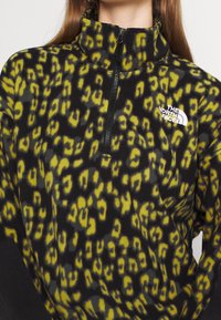 Pullover in pile con colletto alto, caratterizzato da un motivo maculato leopardato nero e giallo e una chiusura a zip. Il logo The North Face è visibile.