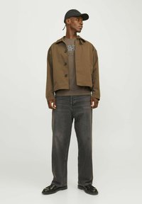 Brun cropped jakke, grå t-shirt med tekst, mørkegrå jeans, sorte sko og en sort kasket. Glat stof, afslappet pasform og subtile detaljer.