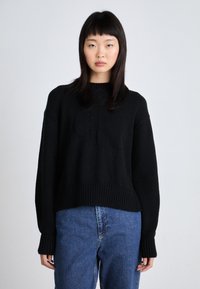 Maglione nero lavorato a maglia con orlo e polsini a coste, caratterizzato da un dettaglio con logo in rilievo. Abbinato a jeans in denim blu, che mostrano una vestibilità comoda.
