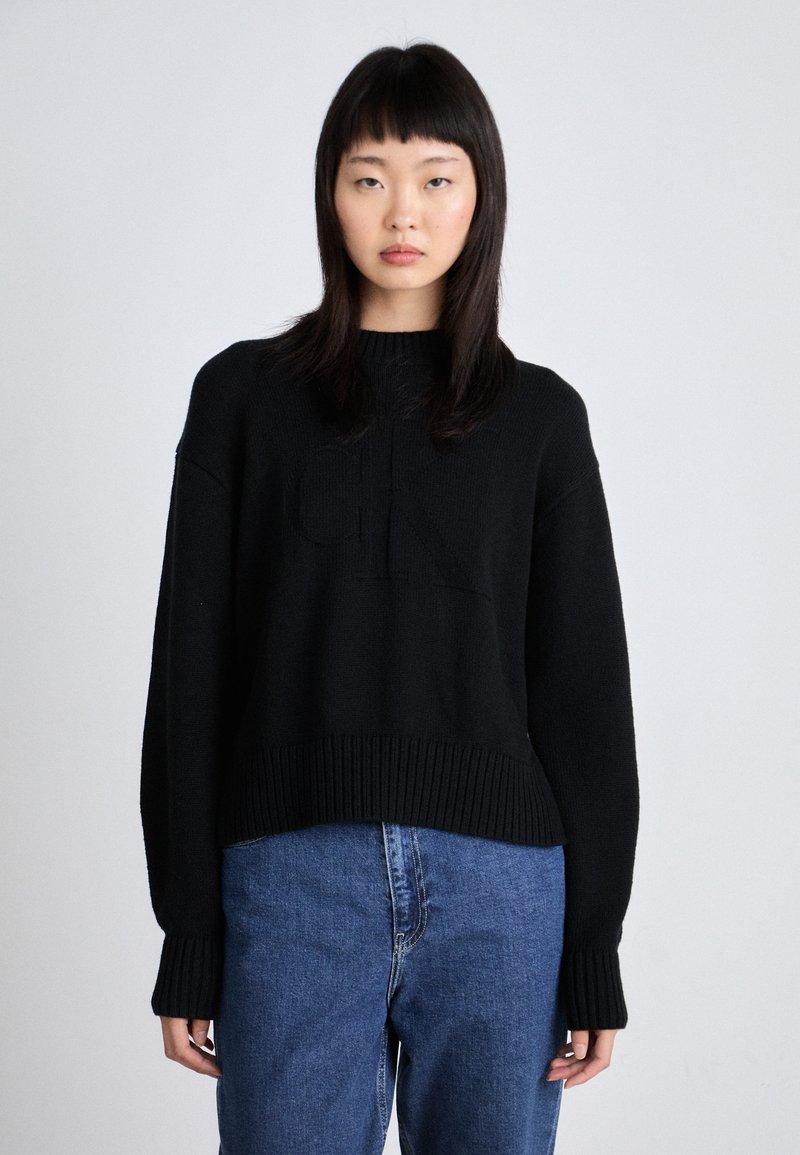 Maglione nero lavorato a maglia con orlo e polsini a coste, caratterizzato da un dettaglio con logo in rilievo. Abbinato a jeans in denim blu, che mostrano una vestibilità comoda.