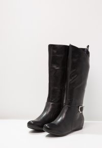 Bottes noires montantes en cuir synthétique lisse. Elles présentent une fermeture éclair sur le côté et une sangle décorative avec une boucle métallique au niveau de la cheville.