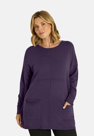 Femme aux cheveux blonds mi-longs portant un long pull violet avec des poches avant et un pantalon noir, debout devant un fond neutre.