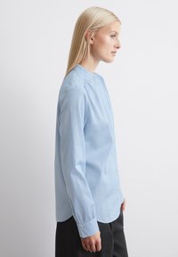 Light blue long-sleeve shirt with a high collar, button front, and subtle pleats at the shoulders. Smooth fabric; relaxed fit.