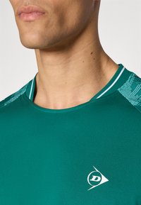 Grünes Sportshirt aus glattem Stoff, mit rundem Kragen und gemusterten Akzenten an den Schultern, sowie einem weißen Logodetail.