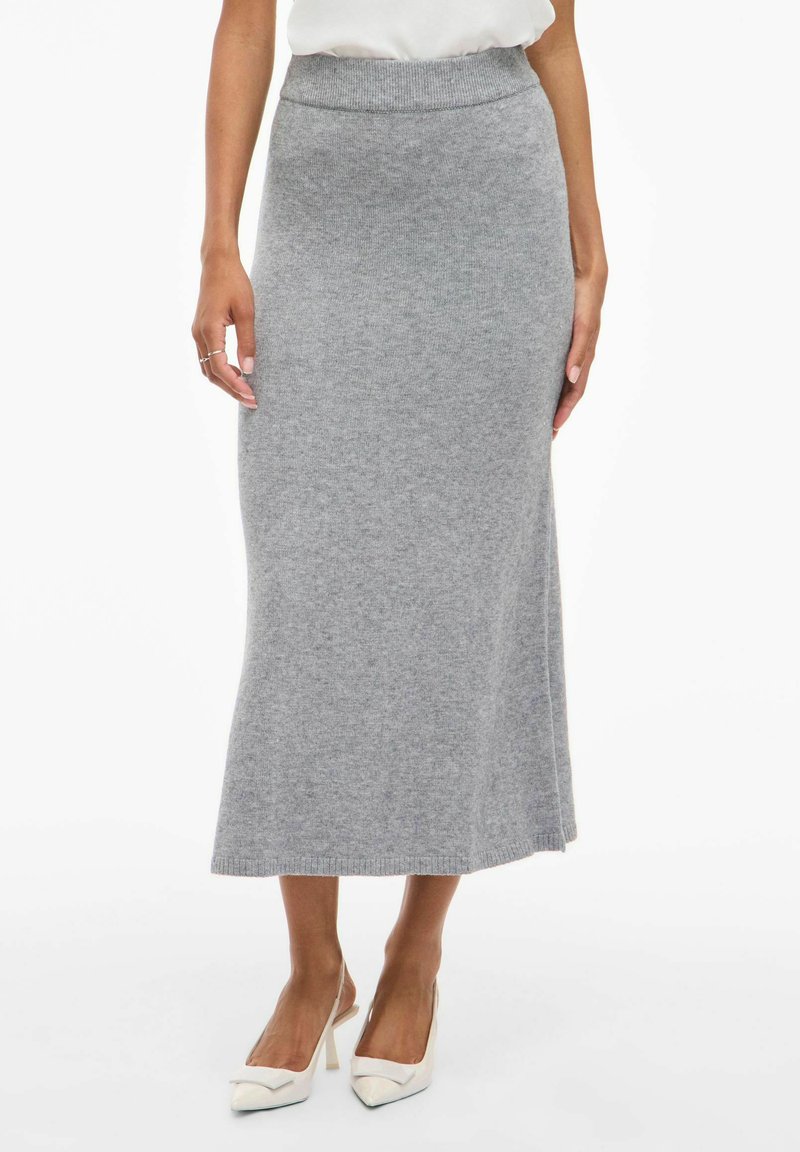 Jupe maxi tricotée grise avec une taille ajustée, texture lisse et légère évasement vers l'ourlet. Portée avec des chaussures à talons hauts blanches.