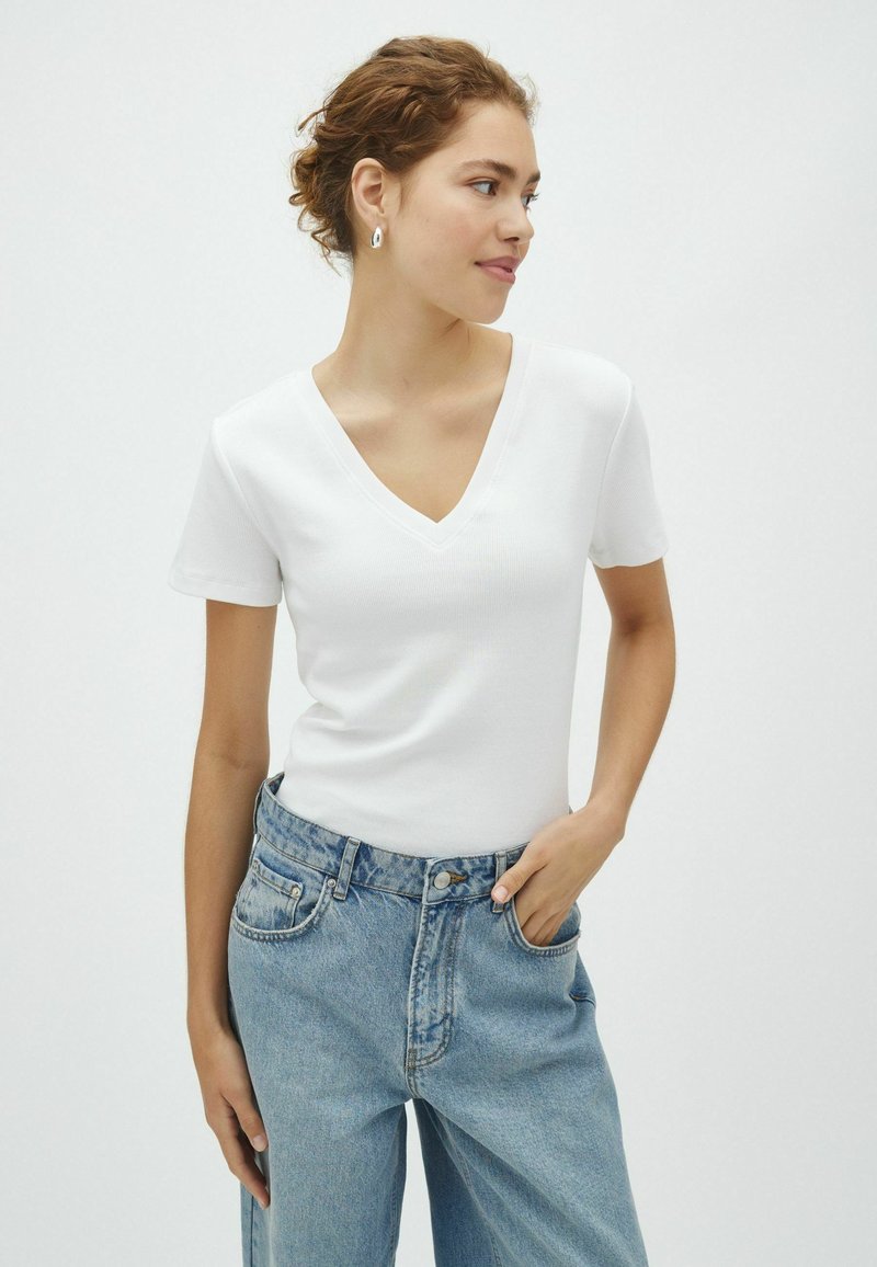 Jeune femme aux cheveux bouclés attachés, portant un t-shirt blanc à col en V et un jean taille haute bleu clair, une main dans la poche.