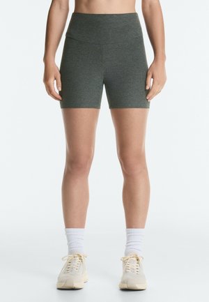 Femme portant un short gris taille haute ajusté, des chaussettes blanches montantes et des baskets beiges à lacets, debout devant un fond uni clair.