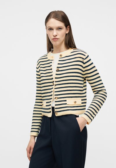 Γυναίκα που φοράει μια μπεζ και ναυτικού μπλε ριγέ ζακέτα με κουμπιά, λευκή μπλούζα και σκούρο παντελόνι, στέκεται με το ένα χέρι στην τσέπη της.