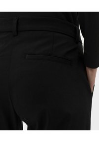 Pantalon noir avec une texture lisse, doté d'une poche arrière unique, d'une fermeture à bouton et de passants de ceinture au niveau de la taille.
