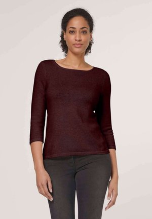 Vrouw met krullend haar, die een donker bordeauxrode top met lange mouwen en een donkere broek draagt, staand tegen een effen lichte achtergrond.
