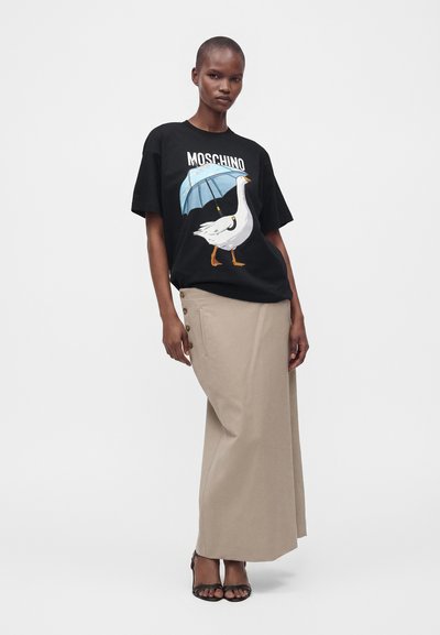 Γυναίκα που στέκεται φορώντας μαύρο t-shirt Moschino με μια χήνα που κρατά μια μπλε ομπρέλα και μια μπεζ μακριά φούστα με κουμπιά στο πλάι.