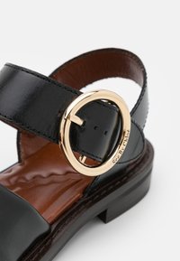 Black leather sandal with a curved strap, featuring a gold circular buckle, brown leather insole, and a low brown heel.
