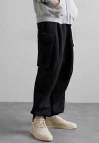 Black jogger pants with two large side pockets, made of soft fabric, featuring a relaxed fit and cuffed ankles. Paired with beige sneakers.