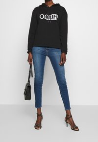 Felpa nera con cappuccio e logo argentato, abbinata a jeans skinny blu e tacchi a strappy neri, con in mano una piccola borsa nera.