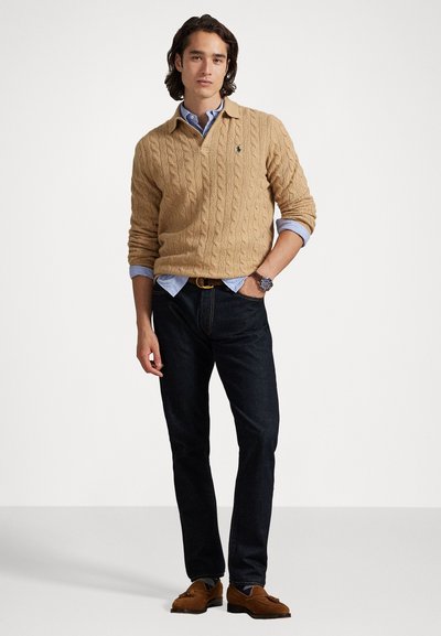 Pull en tricot beige avec un col en V et des manches longues, porté par-dessus une chemise à col bleu clair ; un jean sombre et des mocassins marron complètent l'ensemble.