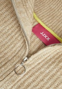 Beige stickad tröja med ett texturerat mönster, med en silverdragkedja och en rosa etikett med gul trim.