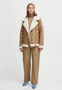 Lichtbruine shearling jas met een witte bontkraag, textuurstof en ritssluiting. Gecombineerd met tan cargobroek en blauwe hoge hakken.