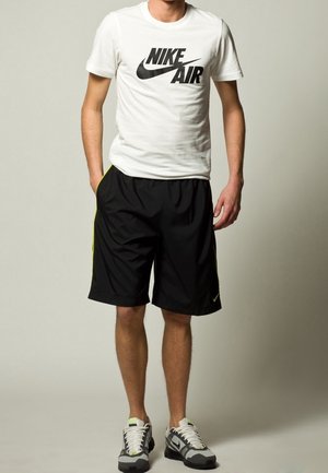 Camiseta blanca de Nike con el logo negro "Nike Air", combinada con pantalones cortos negros que tienen una franja lateral verde y zapatos grises de Nike.
