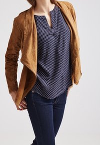 Hnědá semišová bunda s asymetrickým střihem, spárovaná s námořnickou vzorovanou blůzou s bílými mřížkovými liniemi a tmavými džínami.