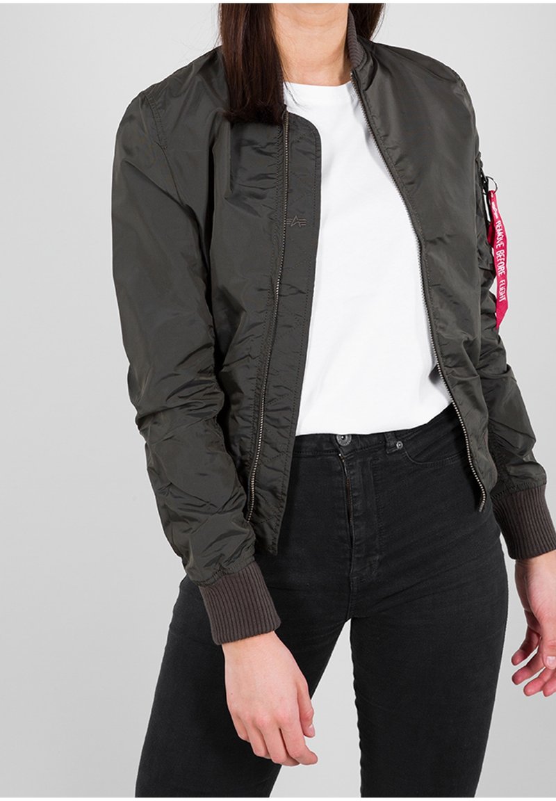 Personne portant une veste bomber vert foncé par-dessus une chemise blanche et un jean noir, debout avec un bras détendu le long du corps.