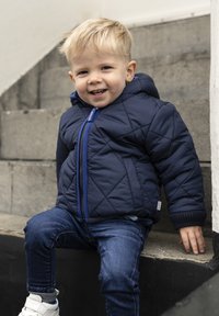 Granatowa pikowana kurtka z kapturem, niebieski zamek błyskawiczny i ściągacze, w połączeniu z niebieskimi dżinsami i białymi trampkami, siedząca na kamiennych schodach.