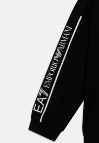 Svart sweatshirtärm med vit text "EA7 EMPORIO ARMANI" och logotyp, samt en kontrasterande vit rand längs kanten. Mjuk tygstruktur.