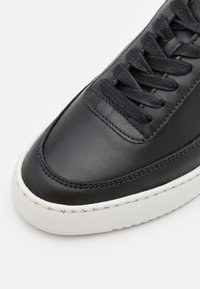 Baskets en cuir noir avec finition lisse, bout rond, semelle plate en caoutchouc blanc et lacets plats noirs, avec des détails de surpiqûres contrastantes.