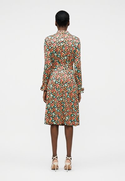 Γυναίκα που στέκεται, φοράει ένα μακρυμάνικο φόρεμα μέχρι το γόνατο με πολύχρωμο λουλουδάτο μοτίβο και μπεζ ψηλοτάκουνα παπούτσια, με την πλάτη γυρισμένη.