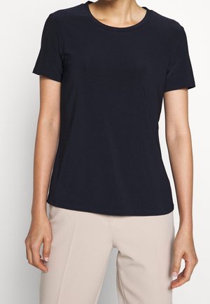 Femme portant un t-shirt noir uni à manches courtes et un pantalon beige clair, debout devant un fond blanc.