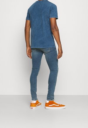 Man draagt een blauw kortärmelig T-shirt, nauwsluitende blauwe jeans en oranje Vans hoge sneakers, staand tegen een effen achtergrond.