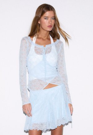 Kobieta o długich włosach nosi przezroczystą, jasnoniebieską koronkową bluzkę z długim rękawem na białym halterze oraz dopasowaną koronkową spódnicę, stojąc na białym tle.