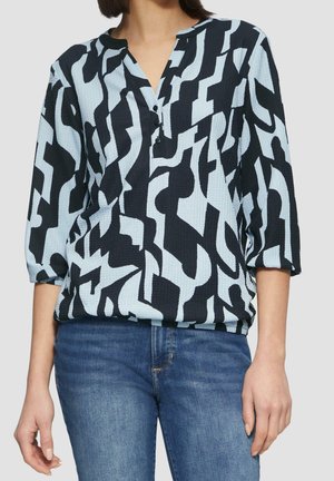 Femme portant un chemisier à motif abstrait noir et bleu clair avec manches trois-quarts et un jean bleu sur un fond clair.