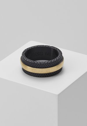 Sort tekstureret ring med en glat guldbånd omkring midten graveret med "GUESS," vist på en hvid pedestal mod en grå baggrund.