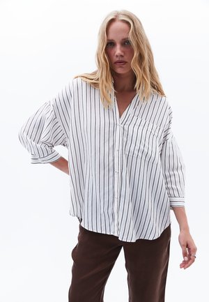 Witte button-up shirt met verticale zwarte strepen, losvallend model, driekwart mouwen, een klein voorzakje, gecombineerd met een paarse broek.