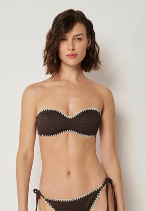 Femme aux cheveux bruns ondulés mi-longs portant un haut de bikini bustier texturé brun foncé et un bas assorti à nouer sur les côtés avec une bordure claire.