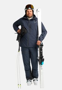 Abbigliamento da sci con una giacca impermeabile blu scuro e pantaloni abbinati. Indossa guanti neri, sci bianchi e scarponi da sci con dettagli turchesi.