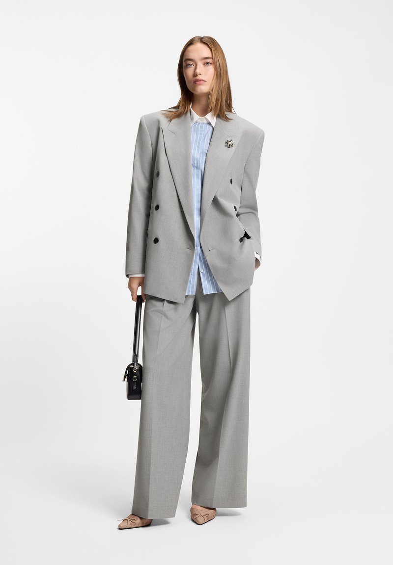 Γκρι σακάκι διπλού στήθους με μαύρα κουμπιά, σε συνδυασμό με ανοιχτό μπλε πουκάμισο με ρίγες, γκρι παντελόνι με φαρδιά μπατζάκια και μπεζ γόβες με μυτερή μύτη.