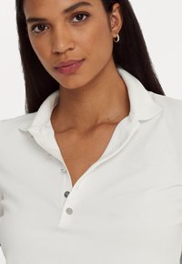 Λευκό πουκάμισο πόλο με κλασικό γιακά, διαθέτοντας κουμπωτή πλέξη, λεία υφή και μικρό λογότυπο κεντημένο στο στήθος.