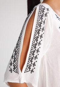 Hvid bluse med korte ærmer med sort blomsterbroderi langs skuldersømmene og udskårne detaljer på ærmerne.