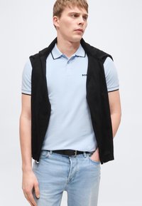 Jeune homme portant un polo bleu clair et un pull noir drapé sur les épaules, debout avec une main dans la poche, regardant sur le côté.