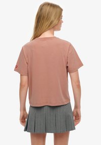 Mujer con cabello largo y rubio, que lleva una camiseta de manga corta rosa polvo y una falda plisada gris con rayas finas, de espaldas sobre un fondo blanco.
