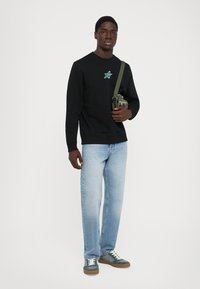 Black sweatshirt with a colorful star design, light blue jeans, and green sneakers. Model holds an olive crossbody bag.